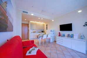 A modern living room with a red sofa, a kitchen area with white cabinets, a dining table with chairs, and a flat-screen TV mounted on the wall. - Appartamento Vista Lago (Desenzano del Garda)