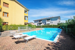 A swimming pool with lounge chairs, a brick patio, and a building with balconies in the background.