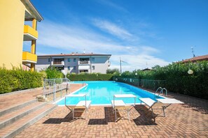 A rectangular swimming pool with a clear blue water surface, surrounded by a brick patio. There are four white lounge chairs aligned along the pool's edge.