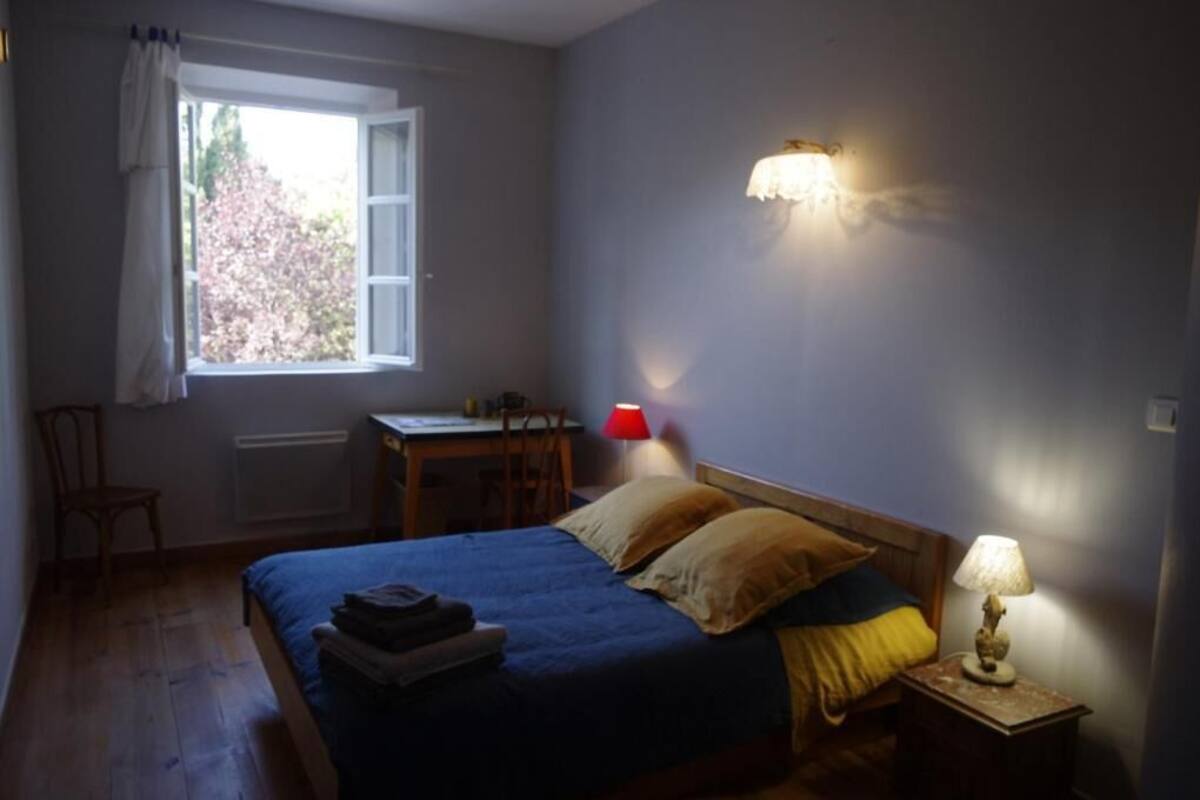 A bedroom with a bed, a desk, a chair, a window with curtains, and a wall-mounted lamp.