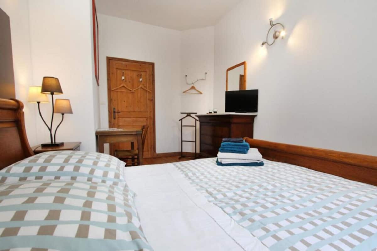 A hotel room with two beds, a wooden wardrobe, a desk with a chair, a television, and a wall-mounted mirror.