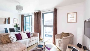 Una sala de estar moderna con un conjunto de sofá, una mesa de centro y una silla. Hay una ventana con cortinas, un candelabro y un cuadro en la pared.