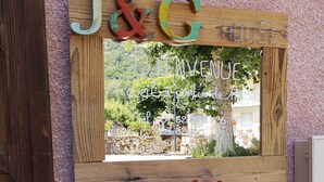 A wooden sign with letters J&G and a welcome message, a mirror reflecting a garden, and a planter with red and white flowers. - J&G House (Saint-Pierre-de-Curtille)