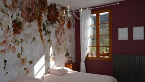 A bedroom with a floral wallpaper, a bed with white bedding, a nightstand with a lamp, and a window with a view of the outdoors. - J&G House (Saint-Pierre-de-Curtille)