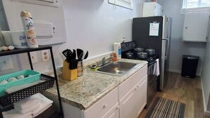 A compact kitchen with a sink, stove, and refrigerator. There is a trash bin and a small table with a chair.