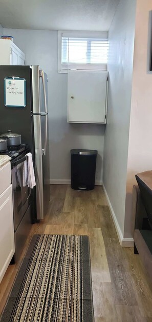 A compact kitchen with a refrigerator, stove, and a trash bin. A welcome sign is on the wall, and a window provides natural light.