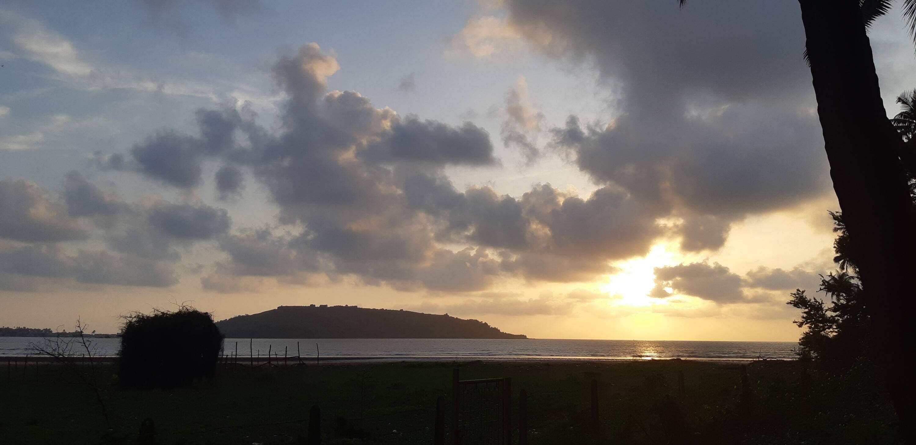 A sunset over a body of water with a distant hill and a fence in the foreground.