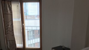 Et soveværelse med seng, et lille bord, en stol, et vindue med rullegardiner og udsigt til bygninger.