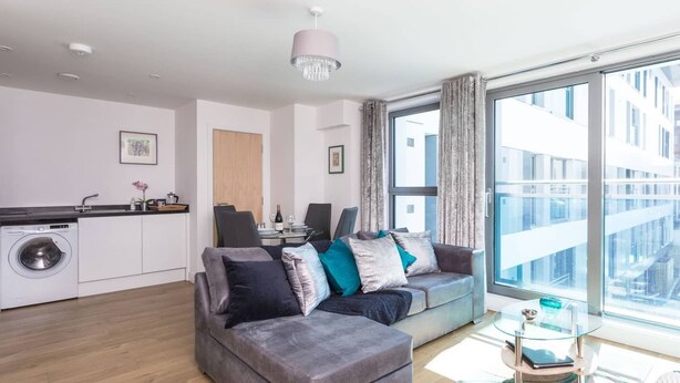 Eine moderne Wohnung mit grauem Sofa, einem Essbereich, einer Waschmaschine und einem großen Fenster mit Stadtblick.