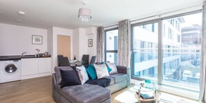 A modern apartment with a grey sofa, a dining area, a washing machine, and a large window with a city view. - Knights Lodge (Redhill)