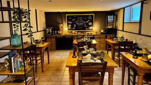 Un coin repas accueillant, avec des tables et des chaises en bois, un tableau noir sur lequel est écrit « BONJOUR », et une cuisine en arrière-plan.