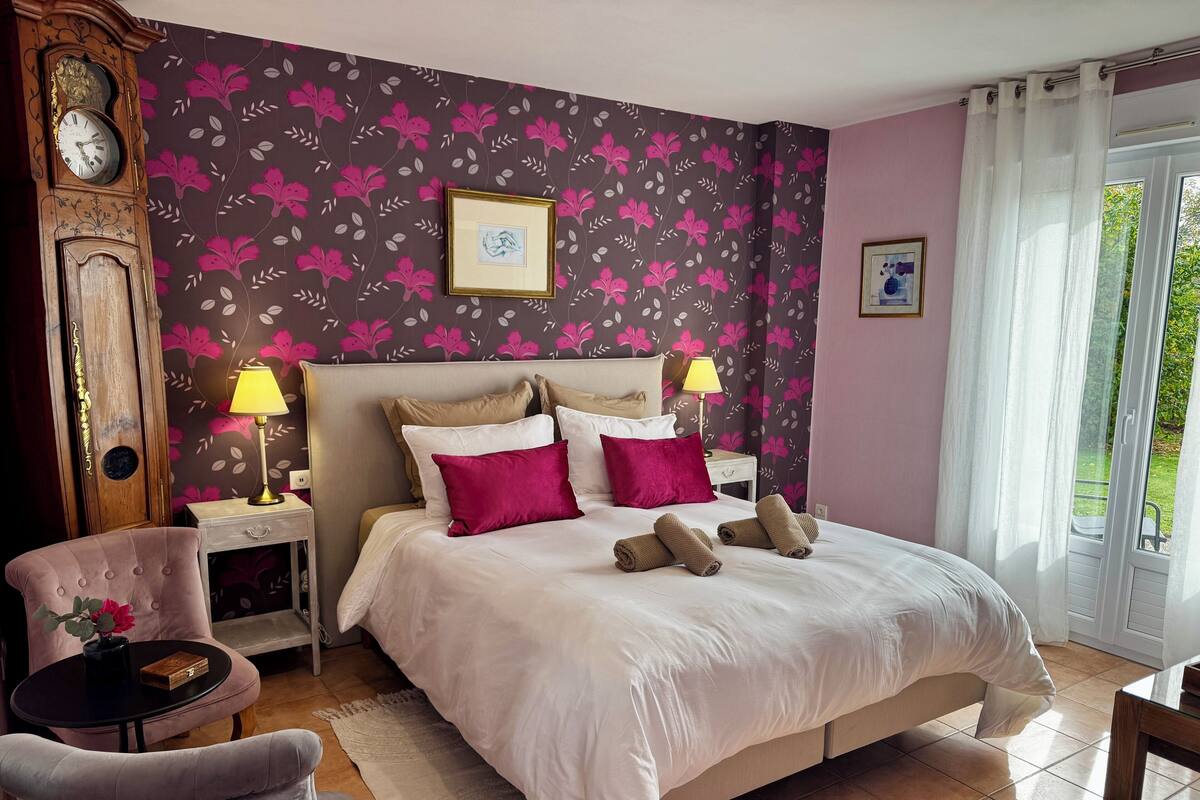Une chambre d’hôtel avec un lit, deux lampes de chevet, une horloge, un tableau encadré et une fenêtre avec des rideaux.