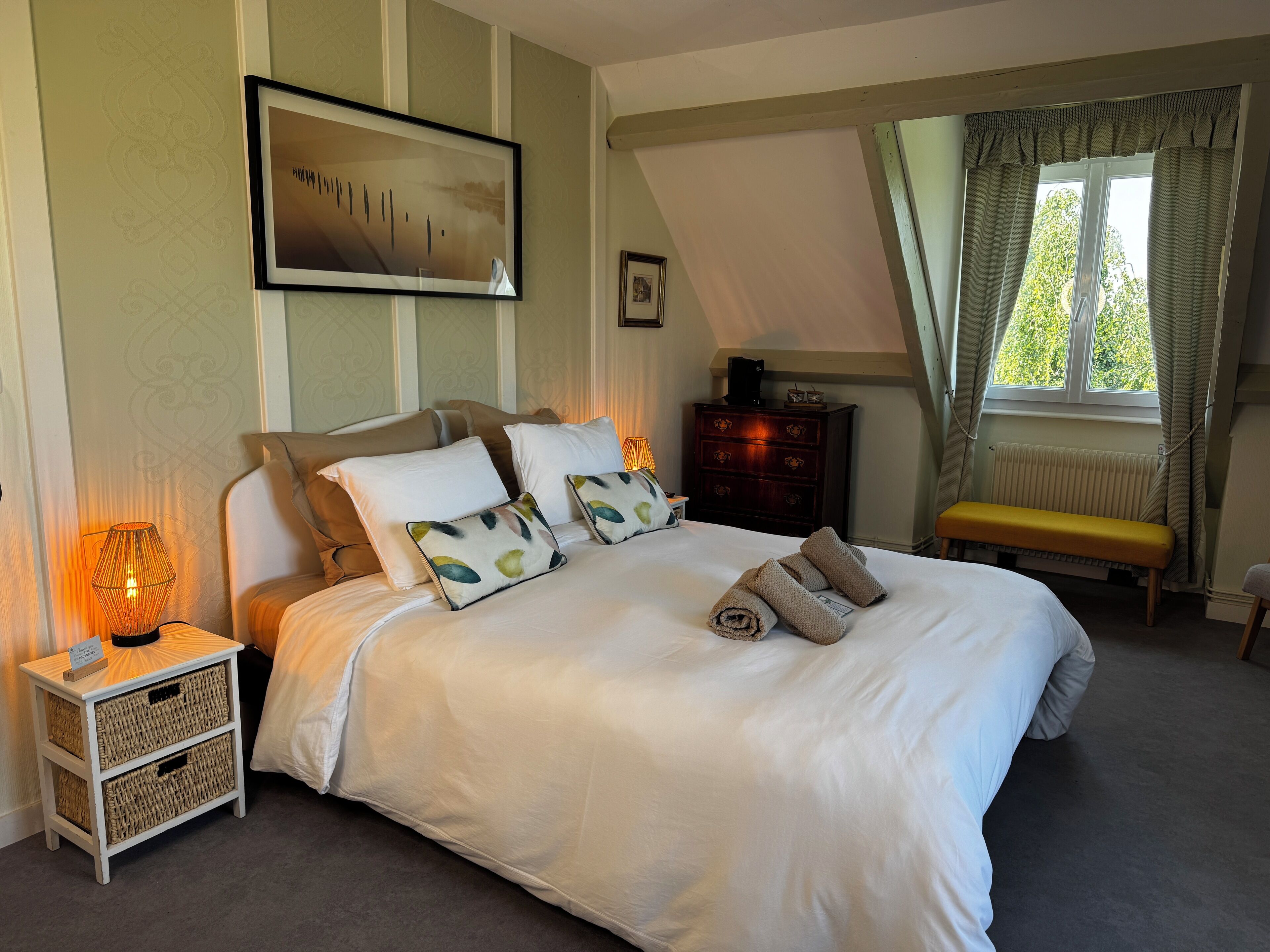A neatly arranged bedroom with a large bed, a bedside table with a lamp, a bench, and a dresser.