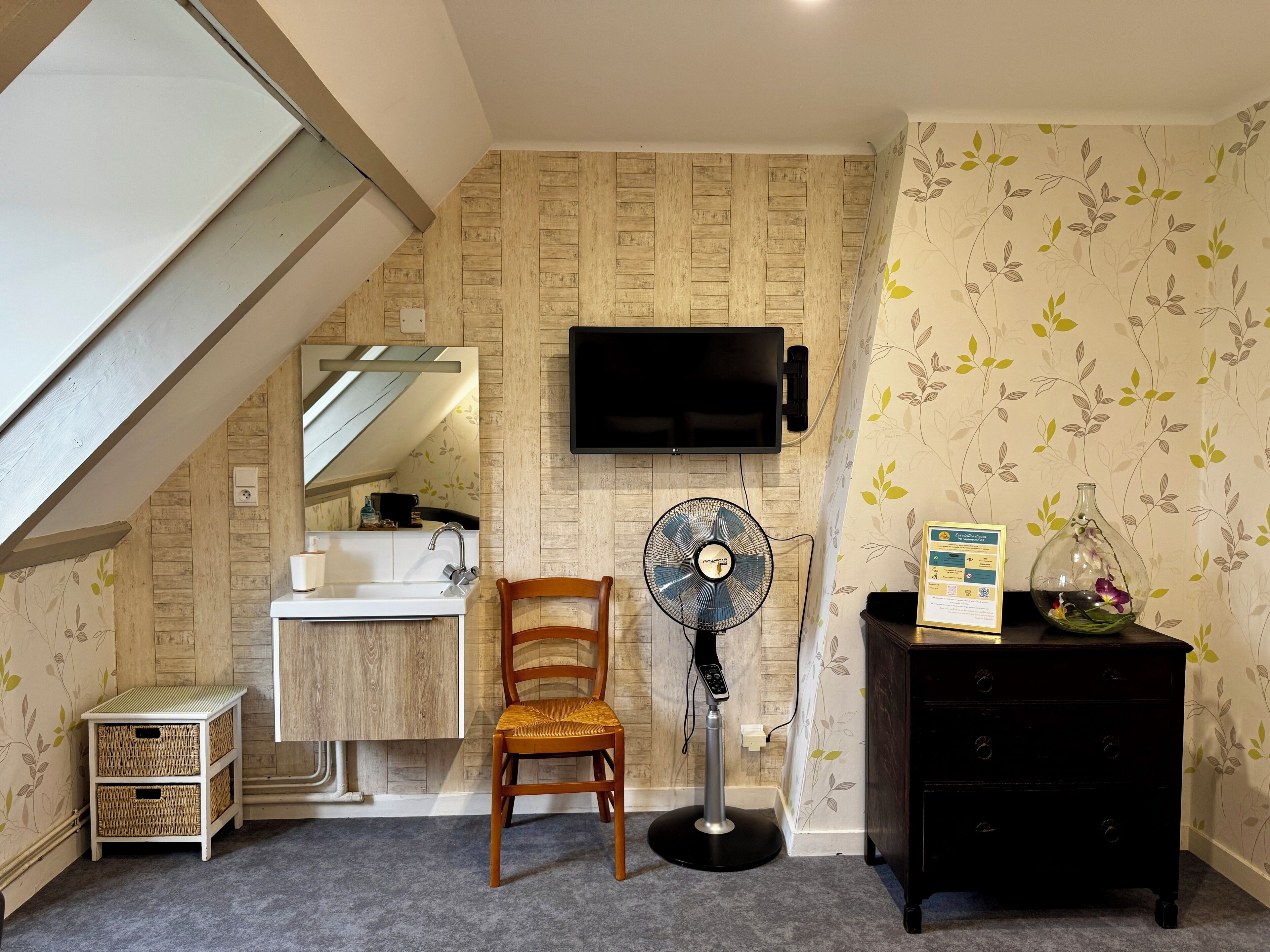 Une salle de bain avec un meuble-lavabo en bois, un miroir, un évier, des toilettes, un ventilateur, une chaise, une commode et un téléviseur fixé au mur.