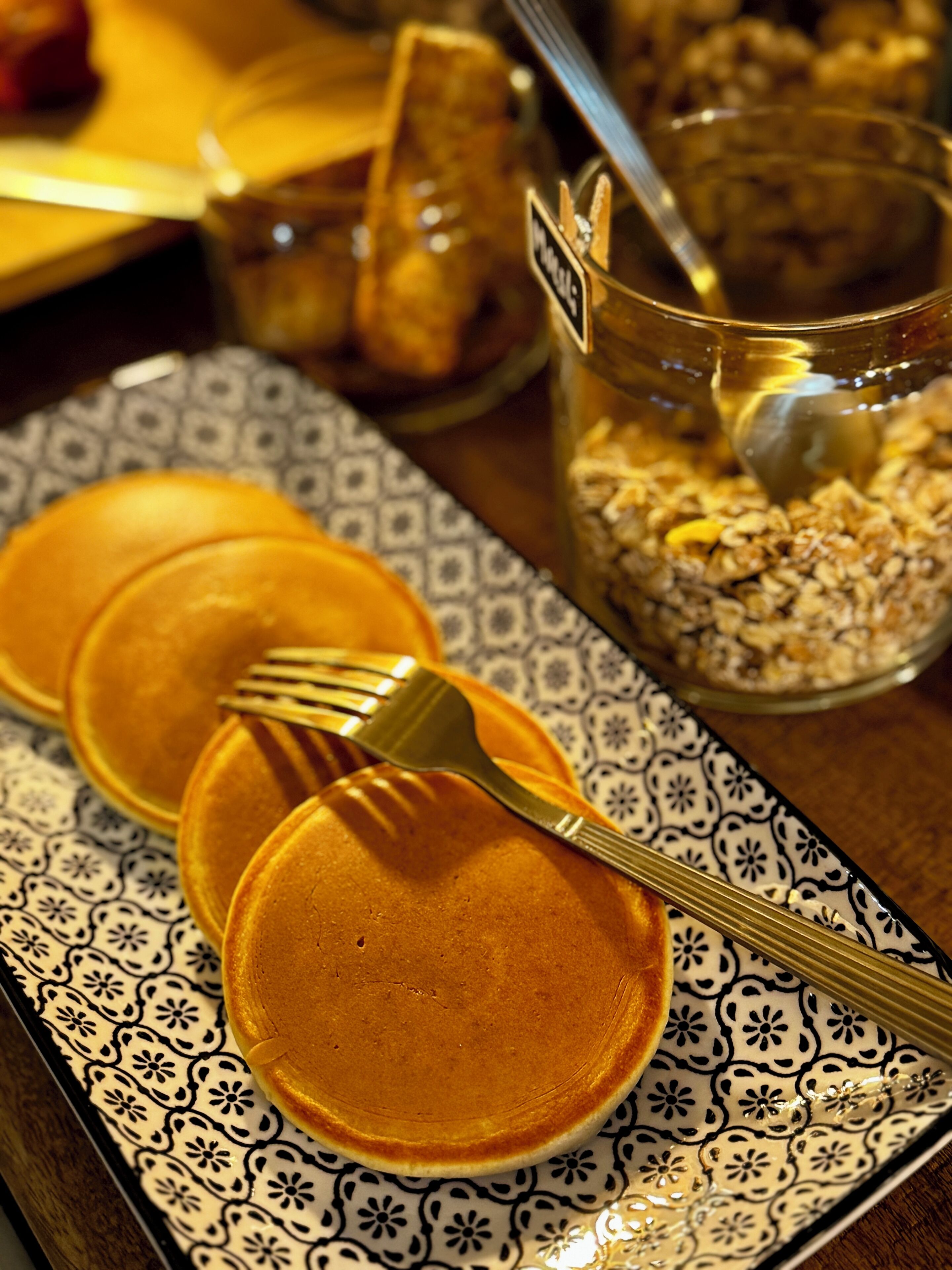 Pancakes on a patterned plate with a fork, a jar of granola, and a spoon.