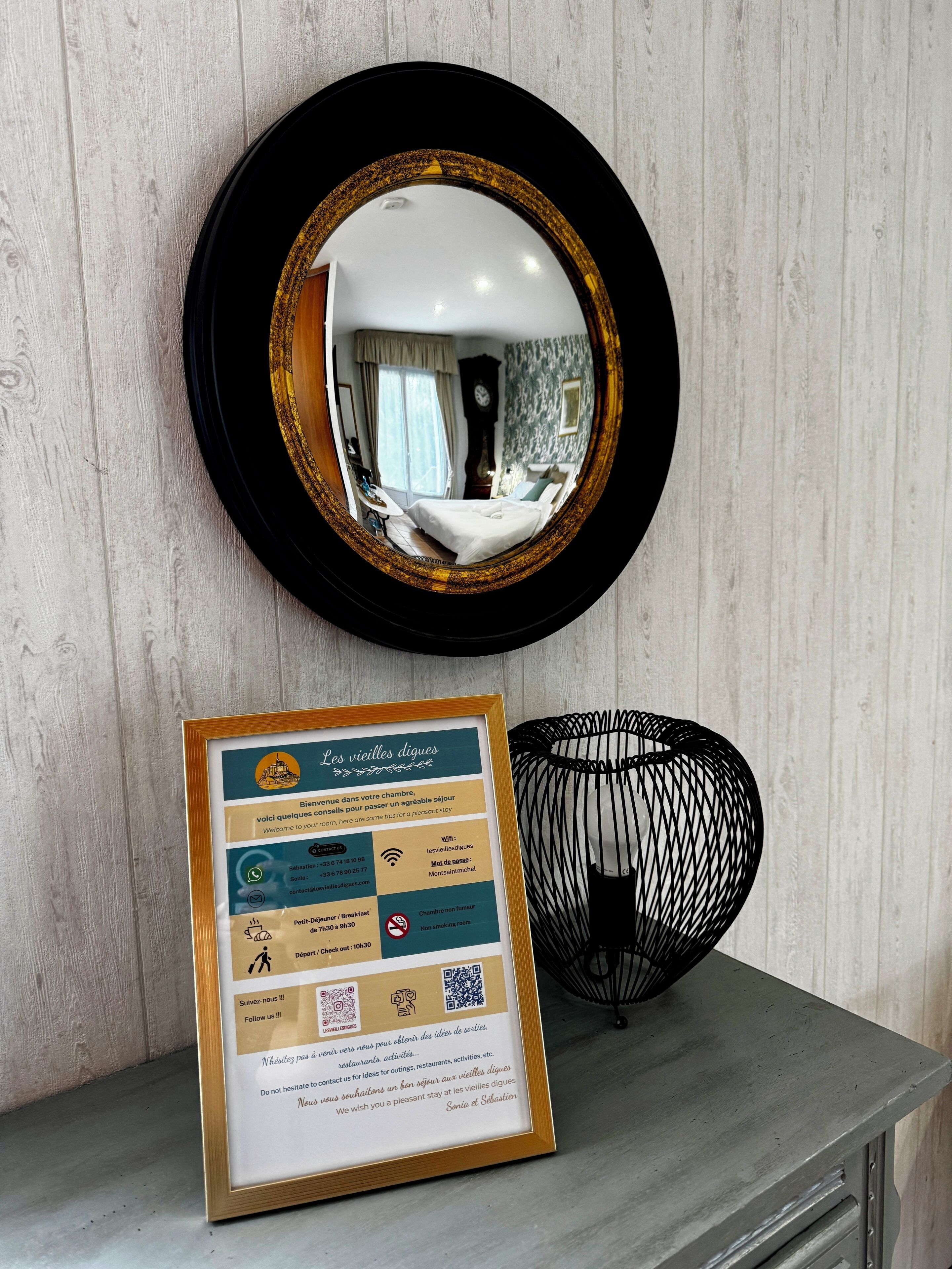Un cartel informativo enmarco con servicios e información de contacto de un hotel, colocado sobre un tocador con espejo y un objeto decorativo.