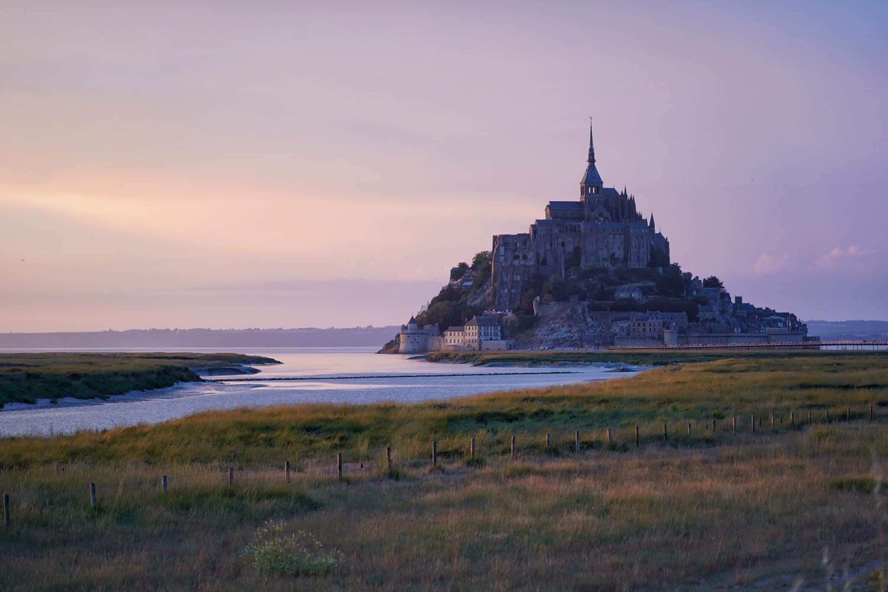 Uma construção em estilo de castelo com uma torre imponente, localizada em uma área gramada e com um corpo d'água ao fundo.