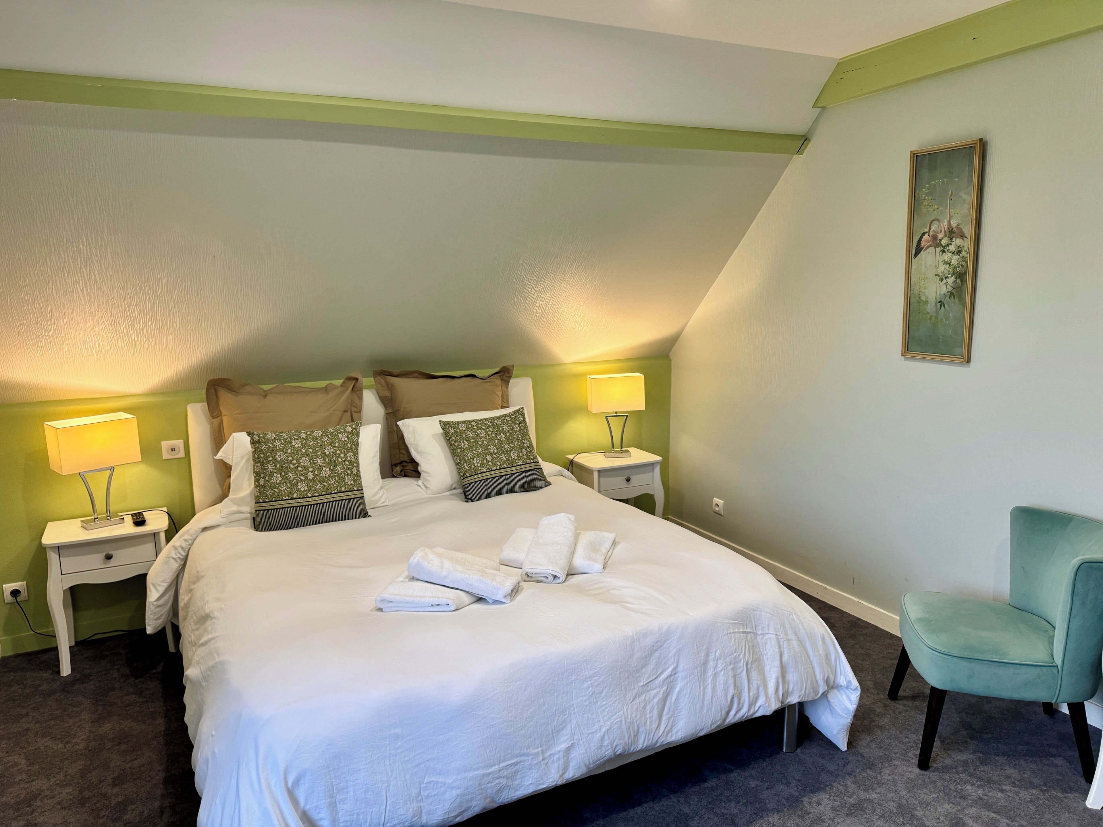 Una cama bien hecha con sábanas blancas y varios almohadones, una silla verde y dos mesitas de noche con lámparas.