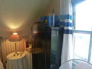 A room with a table lamp, a refrigerator, a fan, and a window with curtains. - Ms McCreadys Guest House (Doncaster)