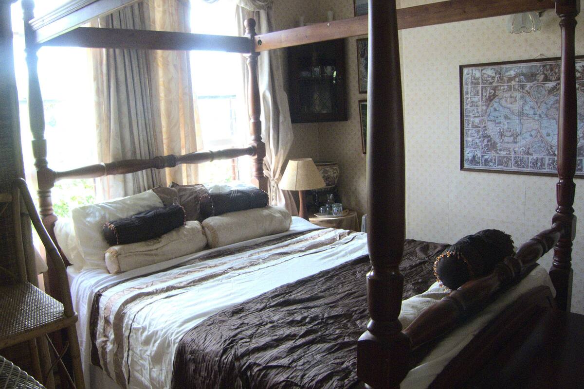 Uma cama de dossel com estrutura de madeira escura, colcha branca e várias almofadas. Há uma pequena mesa com abajur ao lado da cama, e um mapa na parede.