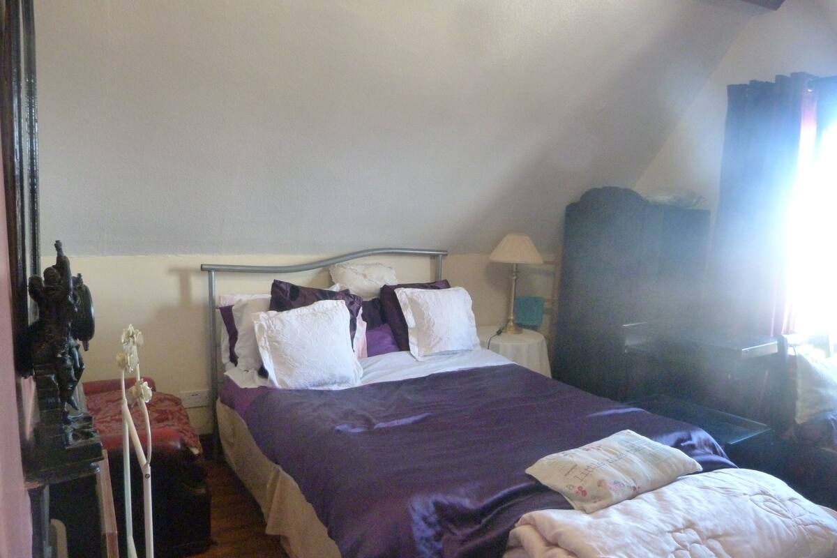 Quarto com cama, almofadas, mesa de cabeceira, abajur e janela com cortinas.