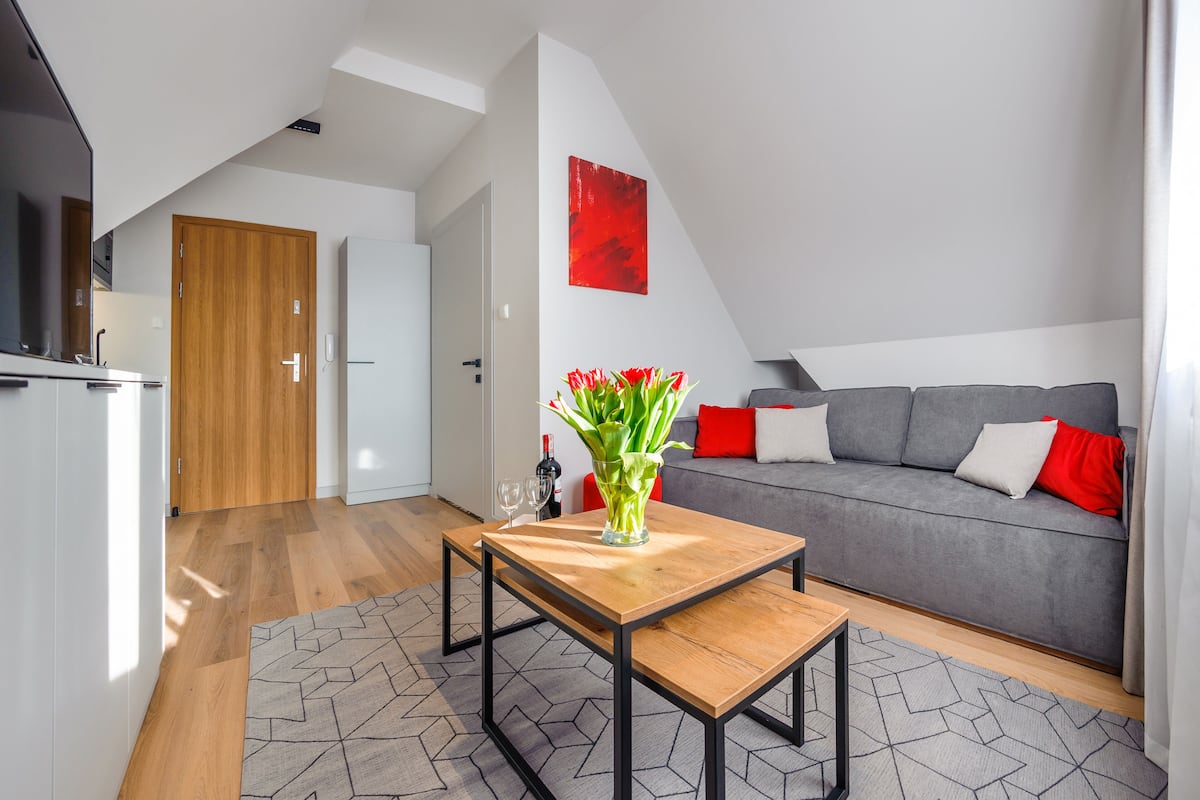 A modern living room with a grey sofa, red cushions, a wooden coffee table, and a vase with red flowers.