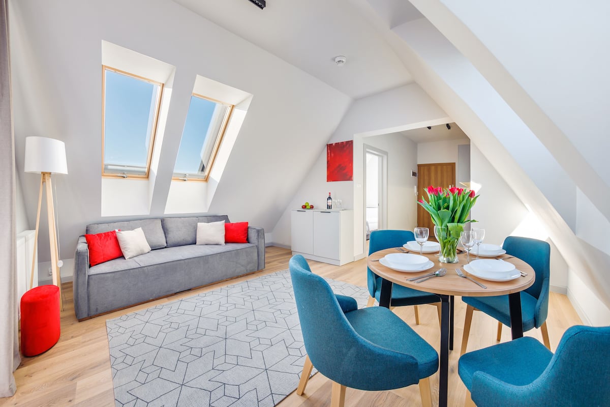 A modern living room with a grey sofa, red cushions, a geometric rug, and a dining area with blue chairs and a wooden table set for four.