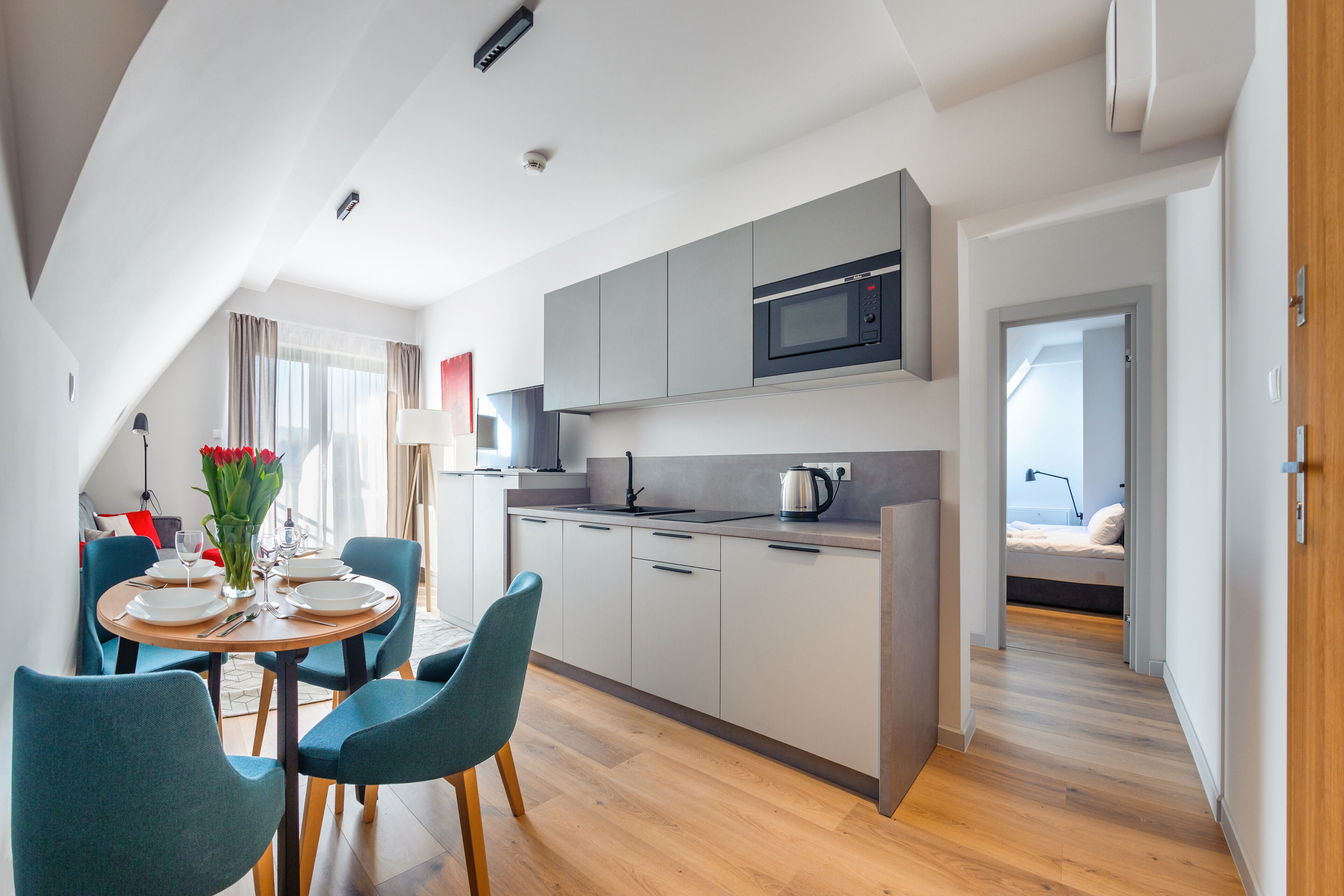 A modern kitchen with grey cabinets, a microwave, and a kettle on the countertop. There is a dining area with a table set for two, and a bed is visible in the background.
