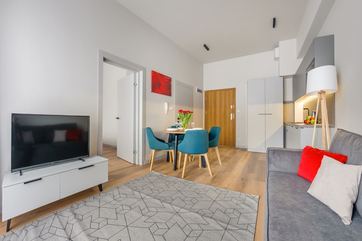 A modern living room with a flat-screen TV, grey sofa, and red cushions. There's a dining area with a table and chairs, and a kitchen area with a lamp.