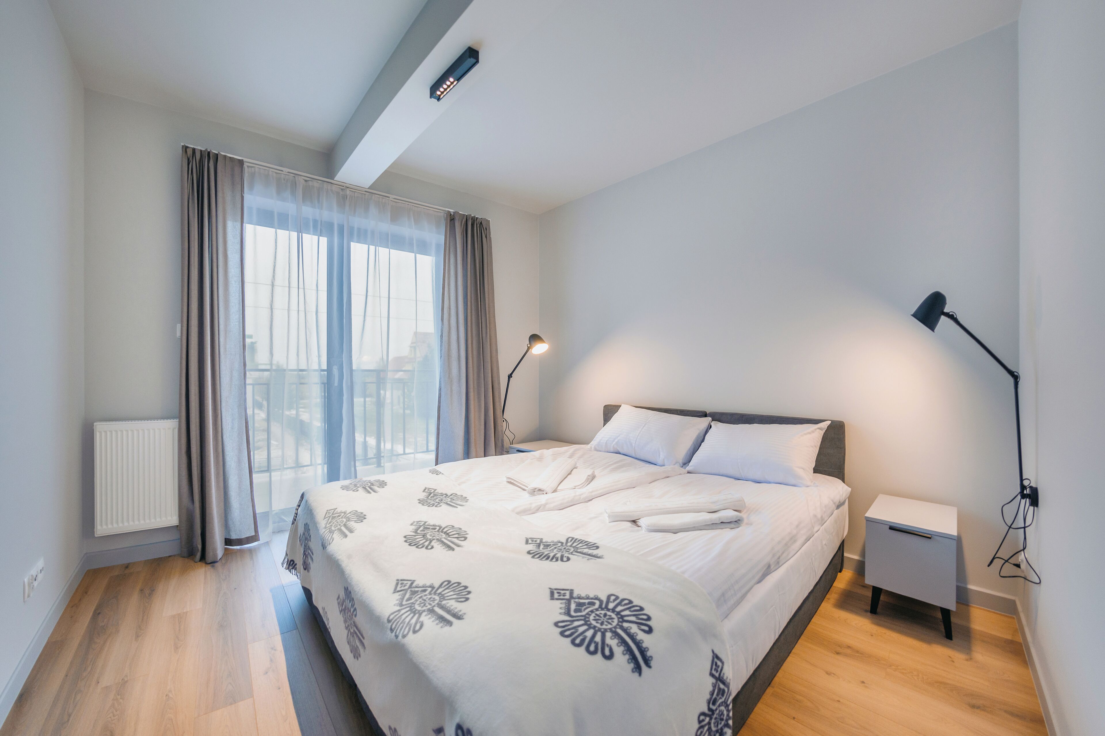 A modern bedroom with a large bed, a bedside table, a lamp, and a view of the city through sheer curtains.