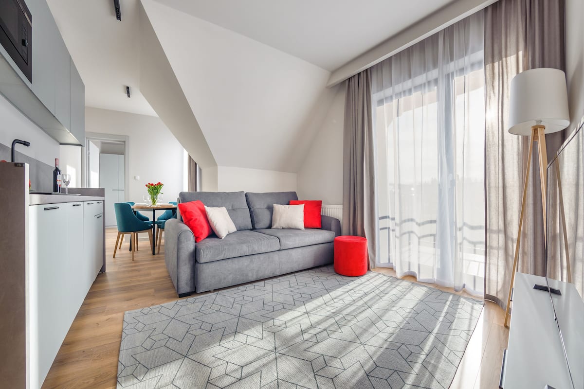 A modern living room with a grey sofa, red cushions, a geometric rug, and a dining area with teal chairs.