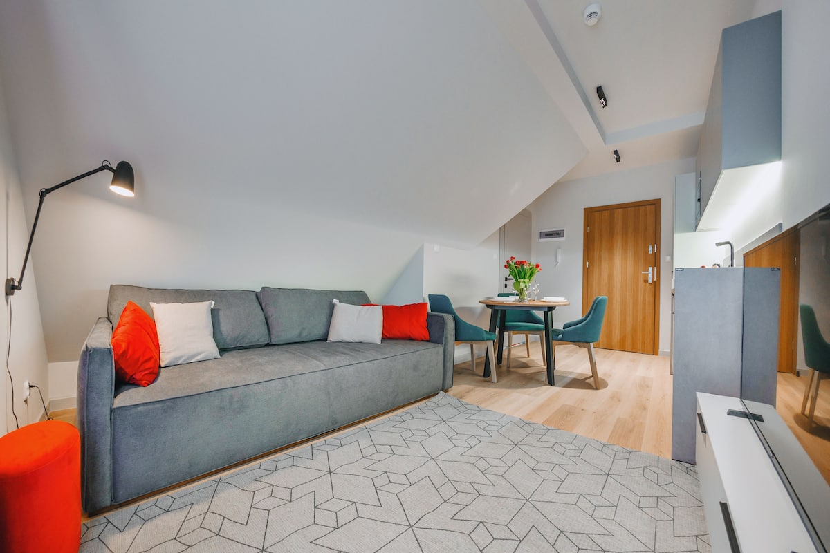 A modern living room with a grey sofa, red and white cushions, a geometric patterned rug, a table with chairs, and a ceiling light.