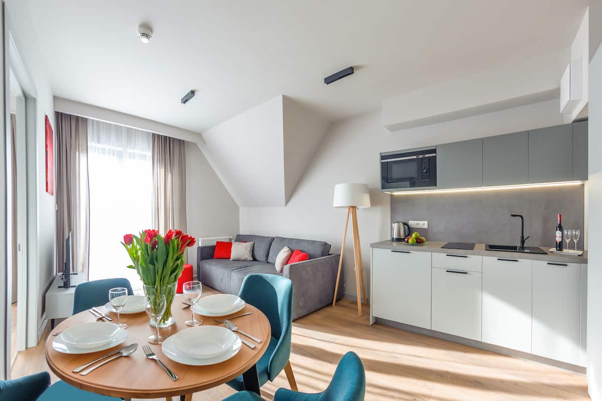 A modern, well-lit room with a dining table set for four, a grey sofa with red cushions, a kitchen area with a microwave, and a floor lamp.