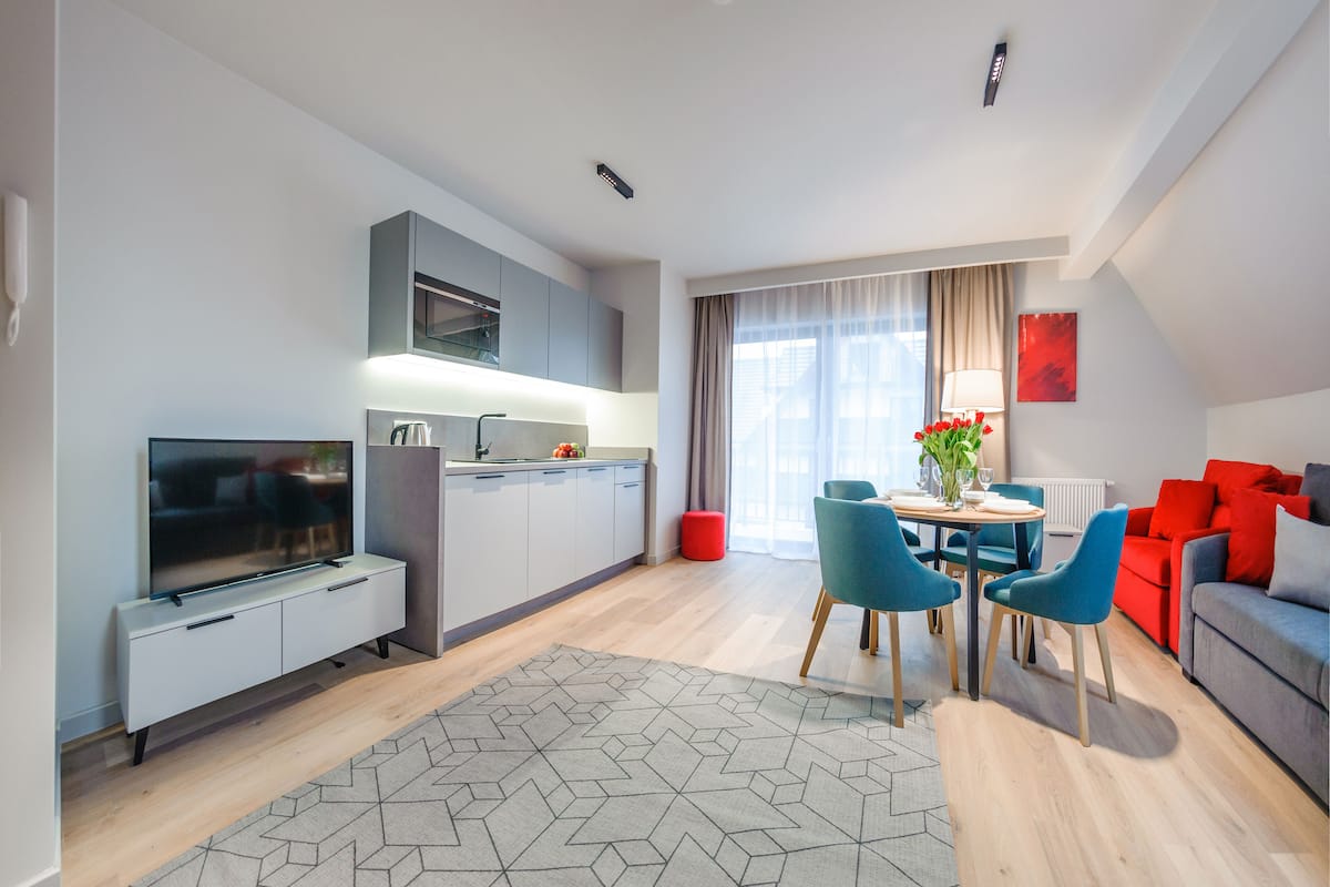 A modern living room with a flat-screen TV, a dining area with a table and chairs, and a sofa with red cushions.