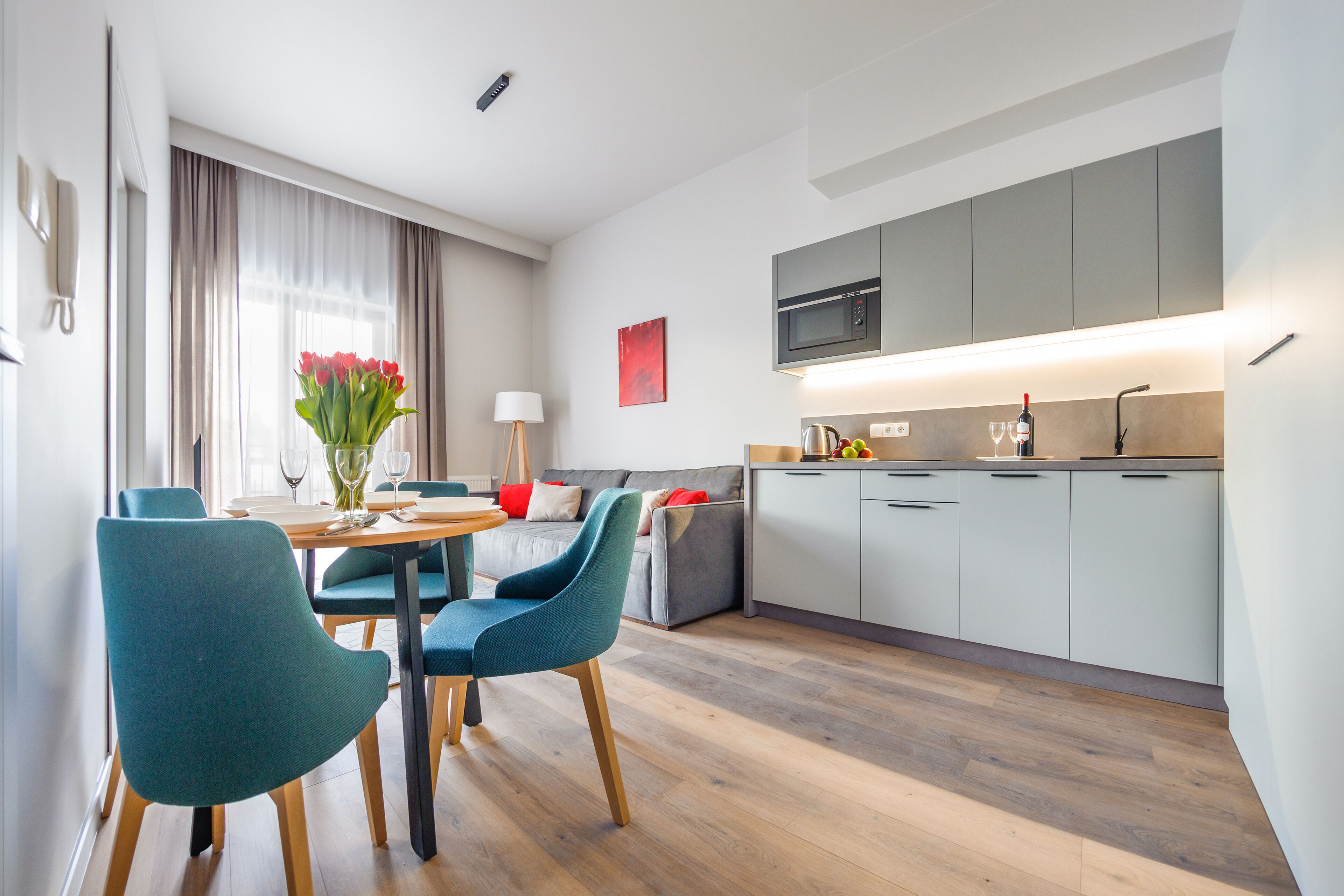 A modern kitchen with grey cabinets, a microwave, and a dining area with teal chairs and a wooden table.
