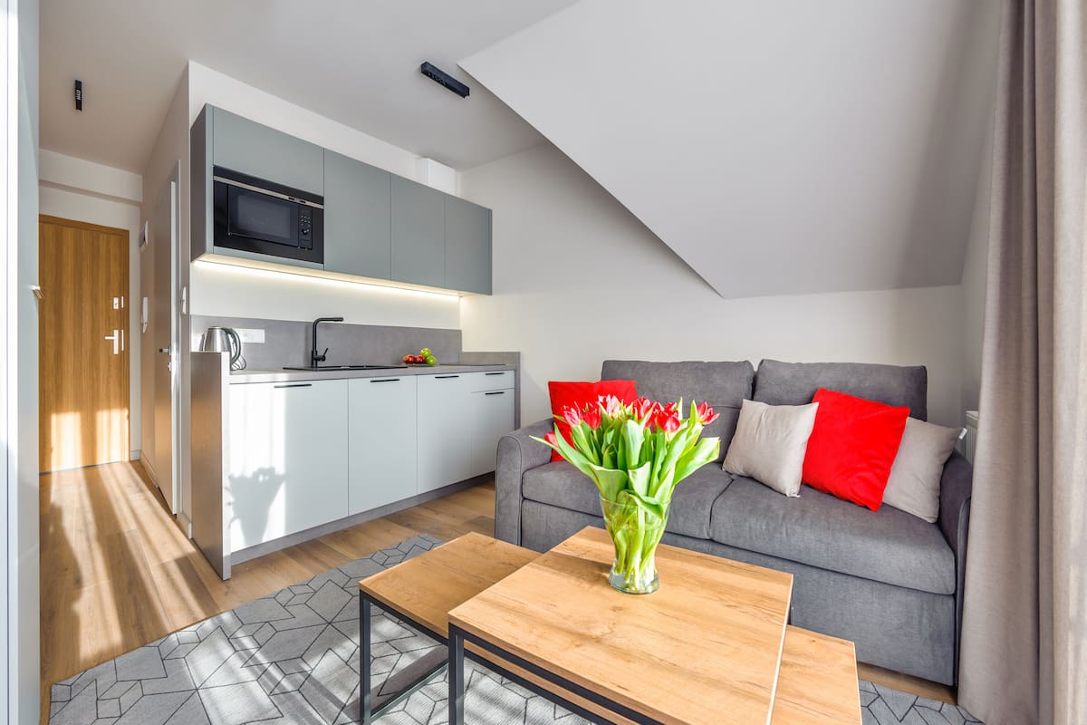 A modern apartment with a kitchen, grey sofa with red cushions, and a wooden coffee table with a vase of flowers.