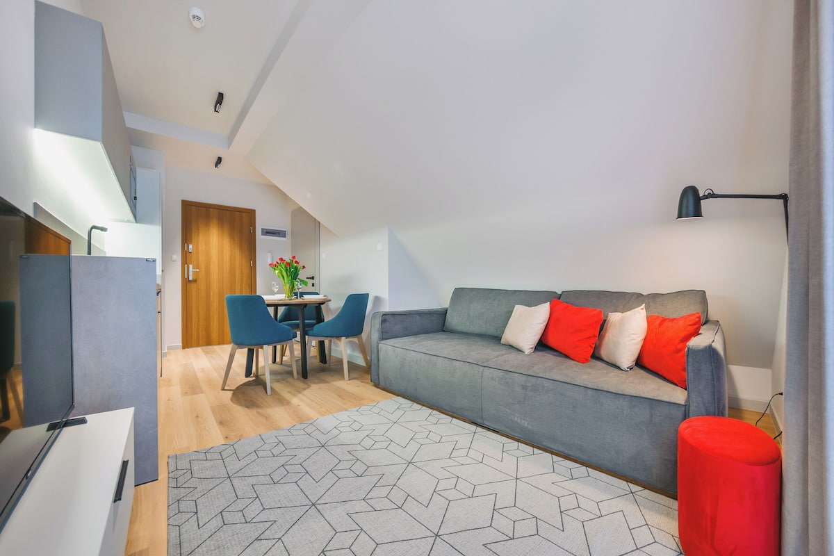 A modern loft apartment with a grey sofa, red cushions, a dining area with blue chairs, and a geometric patterned rug.