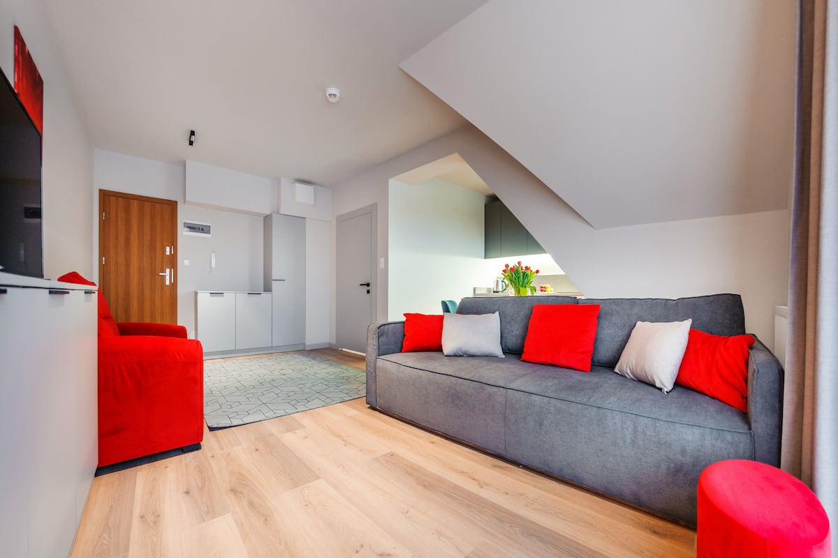 A modern living room with a grey sofa and red cushions, a wooden door, and a kitchen in the background.