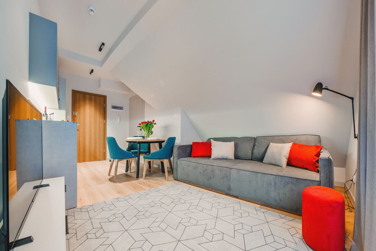 A modern living room with a grey sofa, red and white cushions, a round table with chairs, and a geometric patterned rug.