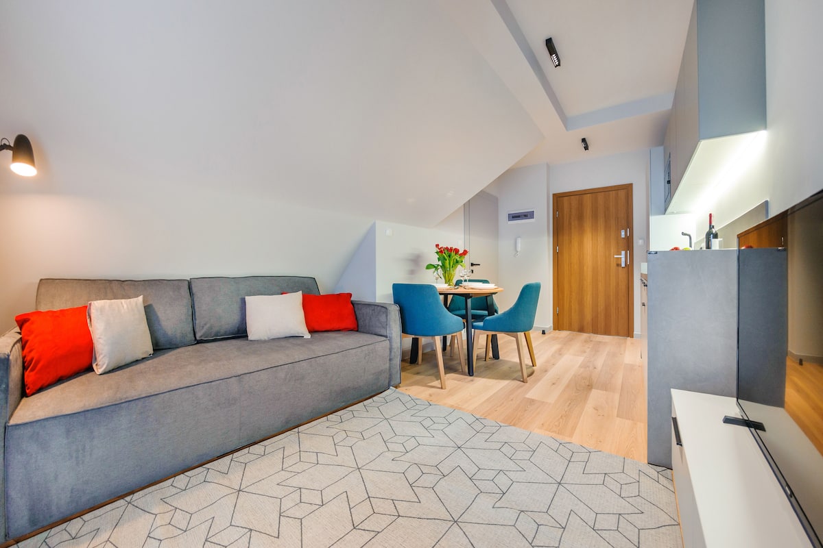 A modern living room with a grey sofa, red and white cushions, a geometric rug, and a dining area with blue chairs and a small table.