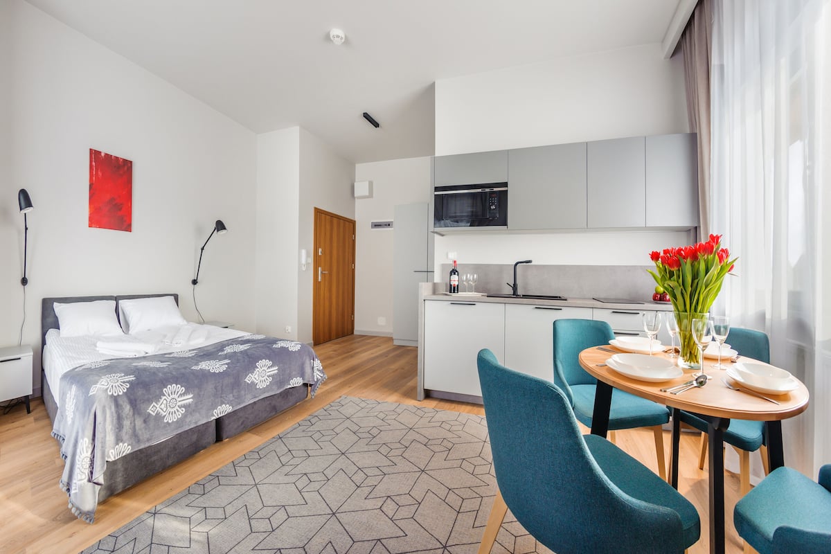 A modern hotel room with a bed, a dining table set for two, a kitchen area, and a red abstract painting on the wall.