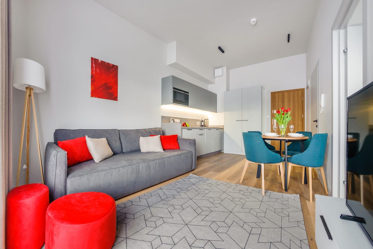 A modern living room with a grey sofa, red cushions, a red ottoman, and a geometric rug. There is a dining area with blue chairs and a table, and a kitchen in the background.