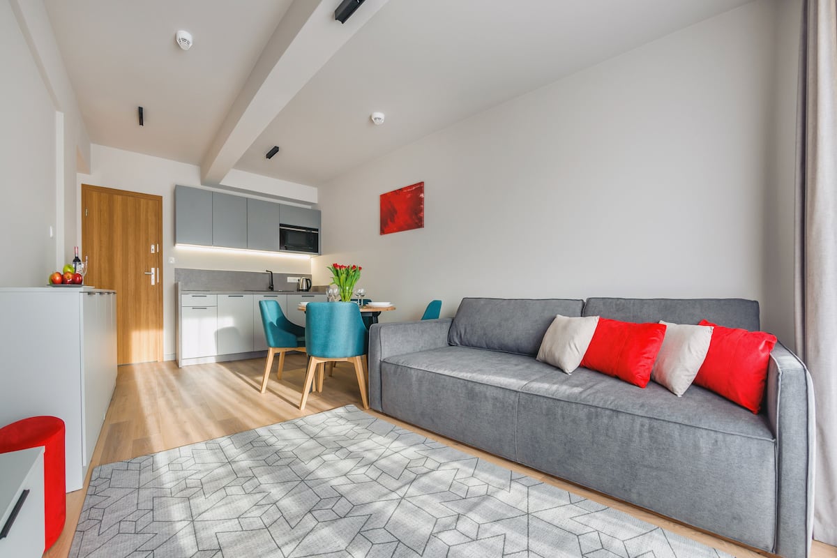 A modern living room with a grey sofa, red and white cushions, a geometric rug, and a dining area with blue chairs and a table.