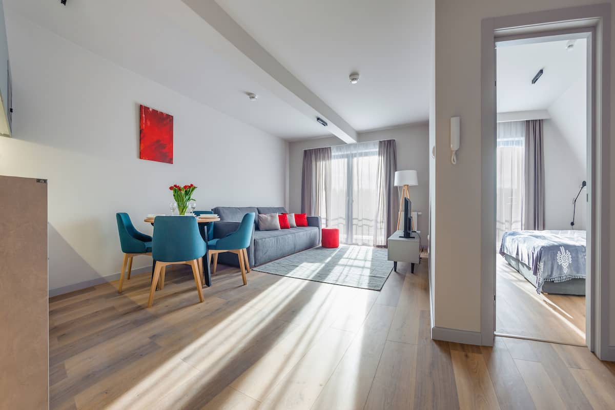 A modern hotel room with a dining area, a sofa with red cushions, and a bed with a blue cover.