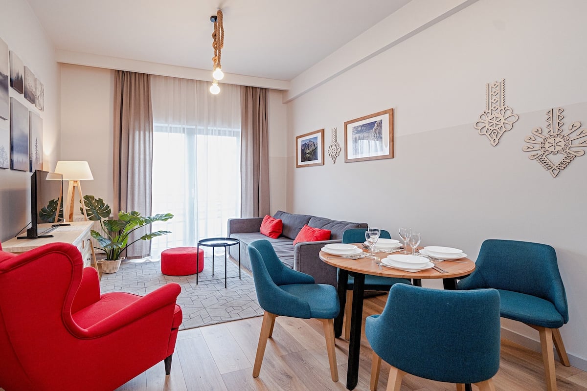 A modern living room with a dining area, red and blue chairs, a sofa, and a large window with curtains.