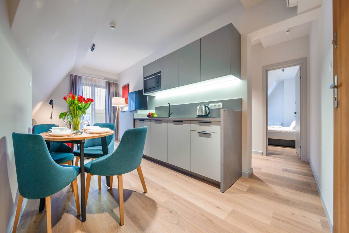 A modern kitchen with grey cabinets, a microwave, and a kettle. There is a dining table with teal chairs and a vase of red flowers.
