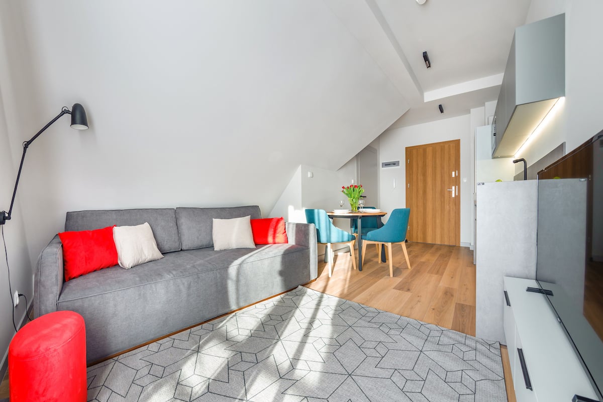A modern apartment with a grey sofa, red cushions, a dining area with blue chairs, and a kitchen with a white fridge.