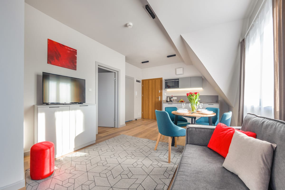 A modern living room with a gray sofa, red and white cushions, and a red ottoman. There is a flat-screen TV mounted on a white cabinet, a dining area with blue chairs, and a kitchen in the background.