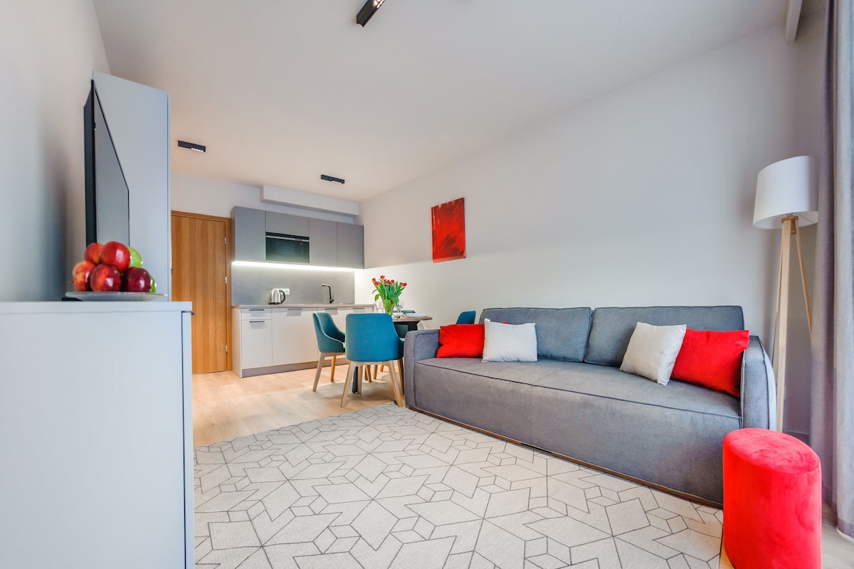 A modern living room with a grey sofa, red and white cushions, a red ottoman, and a geometric patterned rug.