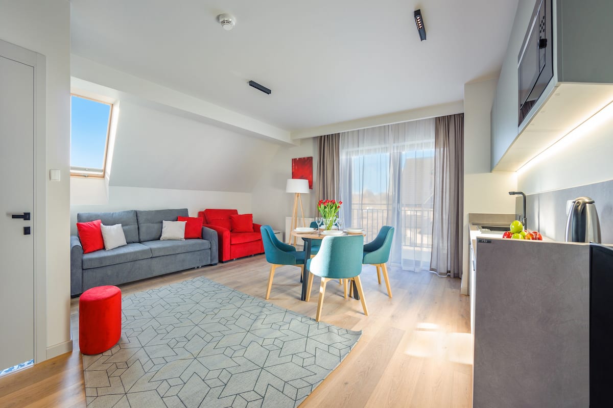 A modern living room with a grey sofa, red cushions, a red ottoman, and a dining area with teal chairs.